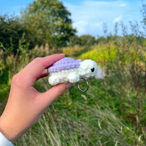 May include: A small crocheted turtle keyring with a purple shell and white body. The keyring is held in a hand against a background of green grass and a blue sky.