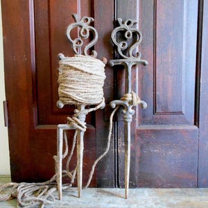 May include: Two antique metal garden twine holders with decorative scrollwork. One holds a large roll of natural twine, while the other is empty. The holders are placed in front of a dark wooden door.
