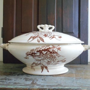 May include: A cream-colored ceramic tureen with a matching lid, featuring brown transferware patterns of birds, foliage, and geometric shapes. The tureen has two side handles and a decorative knob on the lid, resting on a pedestal base.