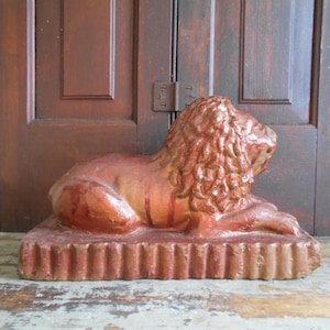 May include: A reddish-brown lion statue in a reclining pose, resting on a rectangular base with vertical ridges. The lion has a detailed mane and is set against a backdrop of dark wooden doors.