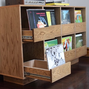 May include: A wooden record player stand with multiple shelves and drawers. The stand is made of light brown wood and has a natural wood finish. The stand has a drawer that is open and a record is visible inside the drawer. The stand has a record player on top of it.