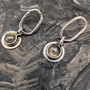 May include: A pair of drop earrings featuring oval-shaped, textured silver hoops connected to circular silver settings. Each setting holds a small, round, translucent green gemstone with visible internal inclusions and a polished finish.