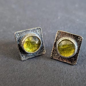 Op de afbeelding: Een paar vierkante oorstekers met een zilverkleurige metalen zetting. Elke oorbel heeft een facetgeslepen, ovaalvormige, geelgroene edelsteen. Het metaal heeft een getextureerd, gehamerd uiterlijk. De oorbellen liggen op een donkergrijze ondergrond.