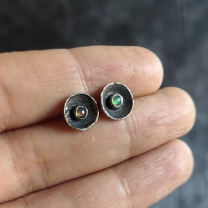 Könnte beinhalten: Ein Paar silberne Ohrringe mit einem kleinen, runden, regenbogenfarbenen Opal-Stein, der in jeden Ohrring eingefasst ist. Die Ohrringe haben die Form einer kleinen, runden Schale.