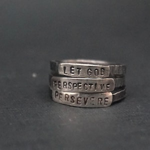 Può includere: Una pila di tre anelli d'argento con le parole "LET GOD", "PERSPECTIVE" e "PERSEVERE" impresse. Gli anelli sono impilati l'uno sull'altro, creando un effetto a strati. Lo sfondo è grigio scuro.