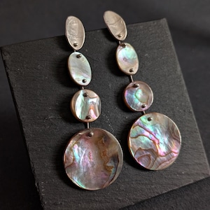 May include: A pair of drop earrings featuring four graduated circular abalone shell discs in each piece. The iridescent shell pieces display swirling patterns of pink, green, and blue, connected by small silver-toned metal links.
