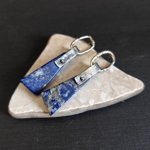 May include: A pair of rectangular lapis lazuli earrings with silver-tone metal accents. The earrings have a dark blue color with gray and white marbling. The silver metal has a textured finish and a small loop for hanging.