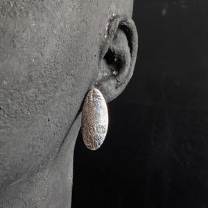 May include: A silver oval earring with a textured surface is shown. The earring is worn on a dark-coloured ear, with the earring's design appearing to be a pattern of small, irregular shapes. The background is a dark, solid colour.