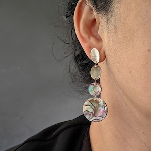 May include: Silver dangle earrings with three iridescent abalone shell discs and a silver oval at the top.