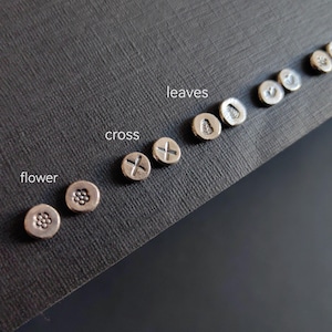 Peut inclure: Un ensemble de boucles d'oreilles en argent avec différents motifs, dont une fleur, une croix et des feuilles. Les boucles d'oreilles sont disposées sur une surface noire avec les mots "flower", "cross" et "leaves" écrits en dessous.