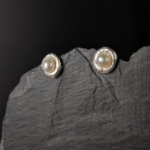 May include: A pair of silver stud earrings, each featuring a lustrous, round pearl. The pearls are set in a textured, silver bezel. The earrings are displayed on a dark, textured surface against a black background.