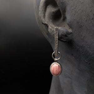 Op de afbeelding: Een zilveren oorbel met een getextureerde staaf en een ronde, roze edelsteen. De edelsteen is gezet in een getextureerde bezel. De oorbel hangt aan een donkergrijze ondergrond, met een zwarte achtergrond.