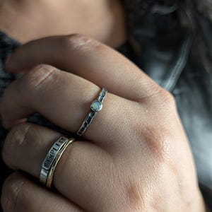 May include: A hand wearing three rings. The top ring is silver with a small, round, opalescent stone. The middle ring is silver with a textured band. The bottom ring is gold with the word "SIMPLE" engraved on it.