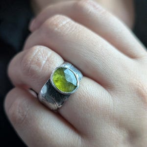 Op de afbeelding: Een zilveren ring met een grote, ovaal geslepen, lichtgroene edelsteen. De ring heeft een band met textuur en bladvormige details. De edelsteen is gezet in een bezel setting.