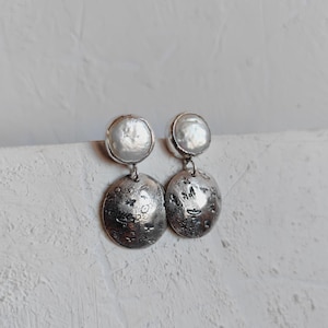 May include: A pair of silver earrings with a textured, hammered finish. Each earring features a round, white pearl at the top and a larger, round, textured silver disc below.