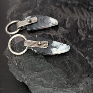 May include: Two handcrafted earrings featuring a black and white stone with silver-toned metal accents. Each earring has a unique pattern and a textured silver loop for attachment. The earrings are displayed on a dark, textured surface.