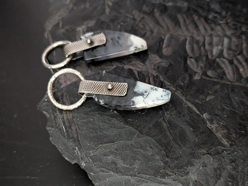 May include: Two handcrafted earrings featuring a black and white stone with silver-toned metal accents. Each earring has a unique pattern and a textured silver loop for attachment. The earrings are displayed on a dark, textured surface.