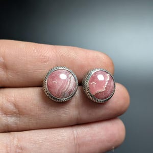 May include: A pair of round stud earrings featuring pink rhodochrosite gemstones set in a silver-toned metal with a textured border. The earrings are displayed against a neutral background.
