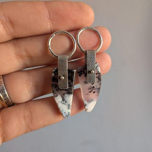 May include: A pair of silver earrings with a unique design. Each earring features a teardrop-shaped stone with black and white patterns, connected to a textured silver band and a circular hoop. The earrings are held up by a hand.