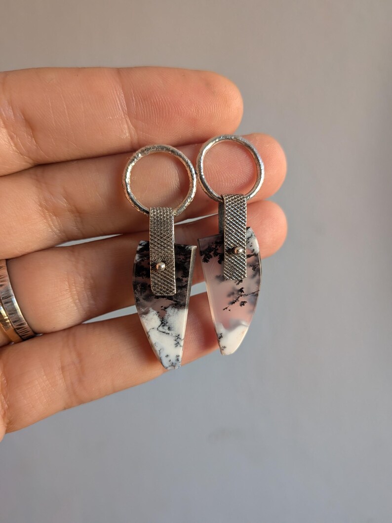 May include: A pair of silver earrings with a unique design. Each earring features a teardrop-shaped stone with black and white patterns, connected to a textured silver band and a circular hoop. The earrings are held up by a hand.