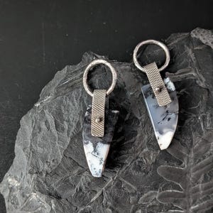 May include: A pair of handcrafted earrings featuring a teardrop-shaped stone with black and white patterns. The stones are attached to a silver-colored metal loop and a textured metal band. The earrings are displayed on a dark, textured surface.