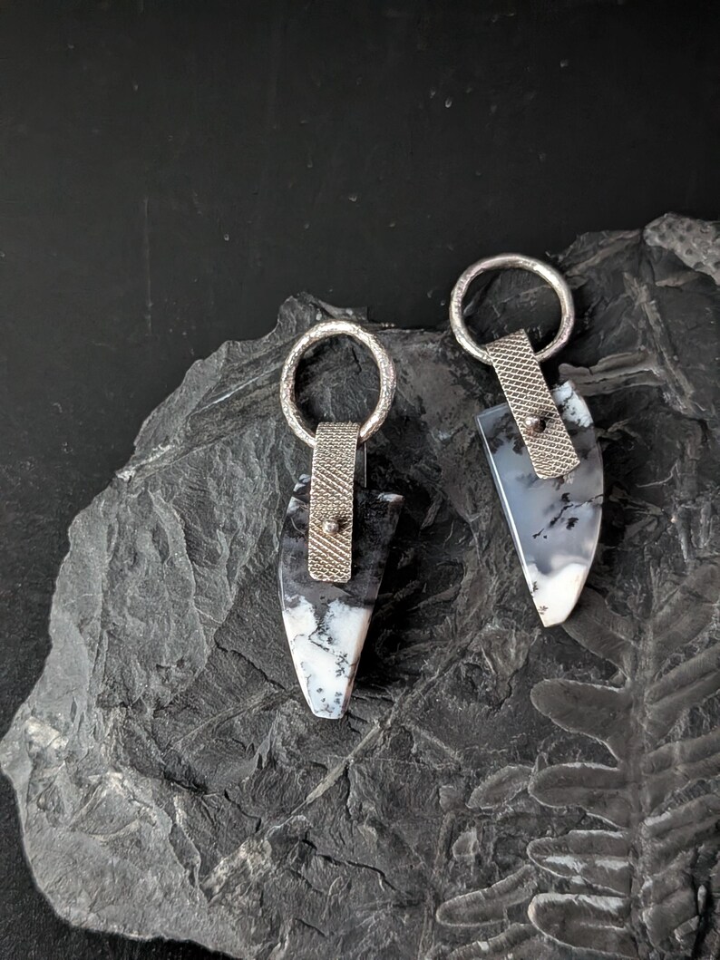 May include: A pair of handcrafted earrings featuring a teardrop-shaped stone with black and white patterns. The stones are attached to a silver-colored metal loop and a textured metal band. The earrings are displayed on a dark, textured surface.