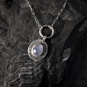 May include: A silver pendant necklace featuring a round, textured metal disc with a central, faceted, light purple stone. The pendant hangs from a silver chain with elongated oval links, showcasing a metallic finish and a detailed, etched surface.
