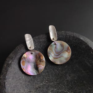 Könnte beinhalten: Ein Paar silberne Ohrringe mit irisierenden Abalone-Muschelakzenten. Die Ohrringe haben eine runde Abalone-Muschel mit einer silbernen Stange, die oben befestigt ist.