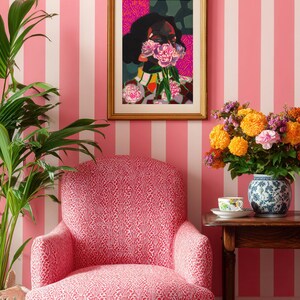 May include: A framed art print featuring a stylized portrait of a person with dark hair holding pink flowers over their face, displayed above a pink and white patterned armchair. The artwork is set in a gold frame against pink and white striped wallpaper.
