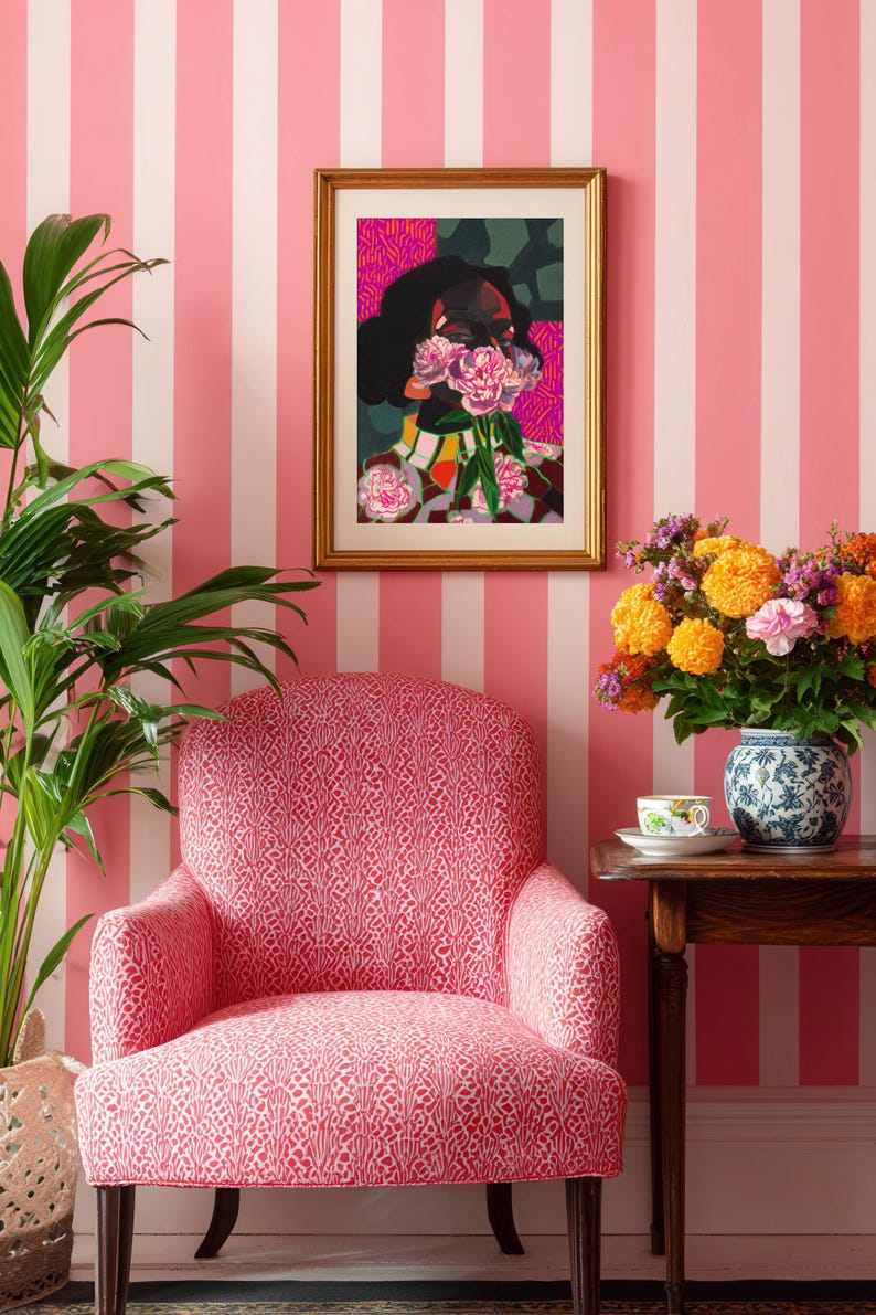 May include: A framed art print featuring a stylized portrait of a person with dark hair holding pink flowers over their face, displayed above a pink and white patterned armchair. The artwork is set in a gold frame against pink and white striped wallpaper.