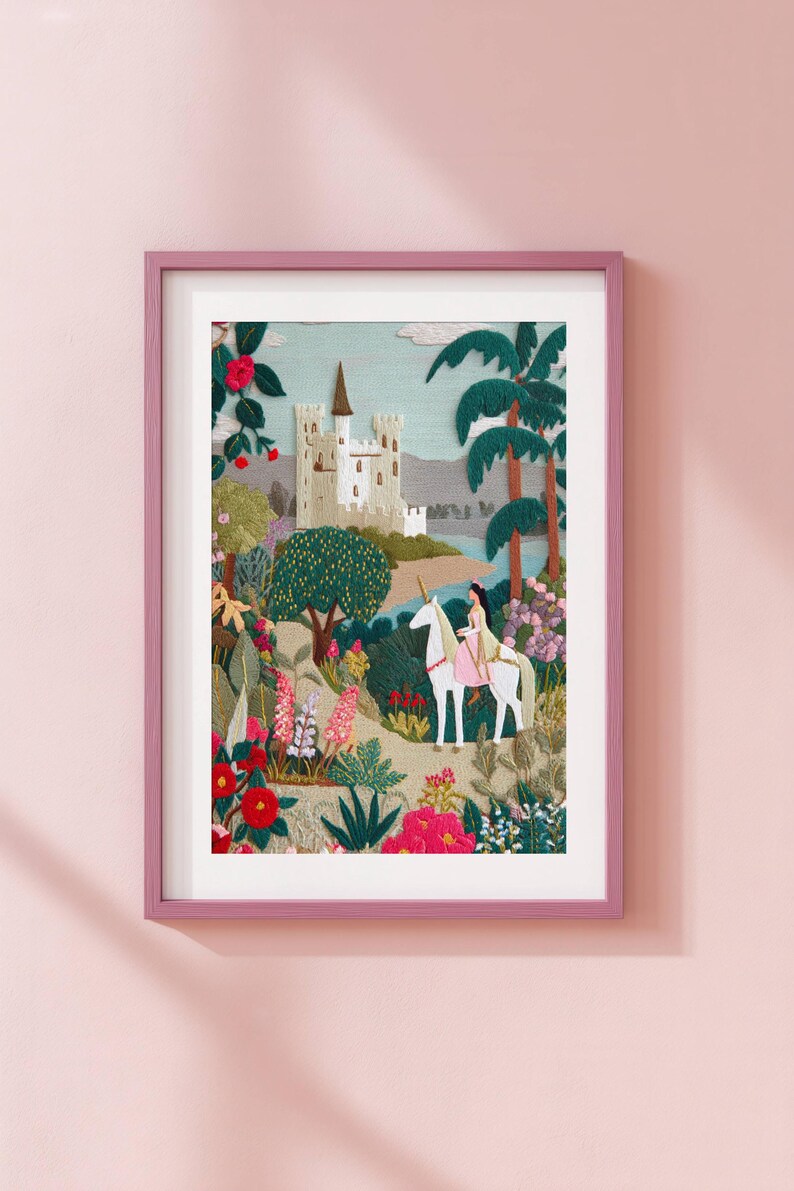 May include: Embroidered artwork in a pink frame depicts a fairytale scene. A woman rides a white unicorn through a colorful garden toward a castle. Palm trees and flowers surround the castle, creating a whimsical landscape.