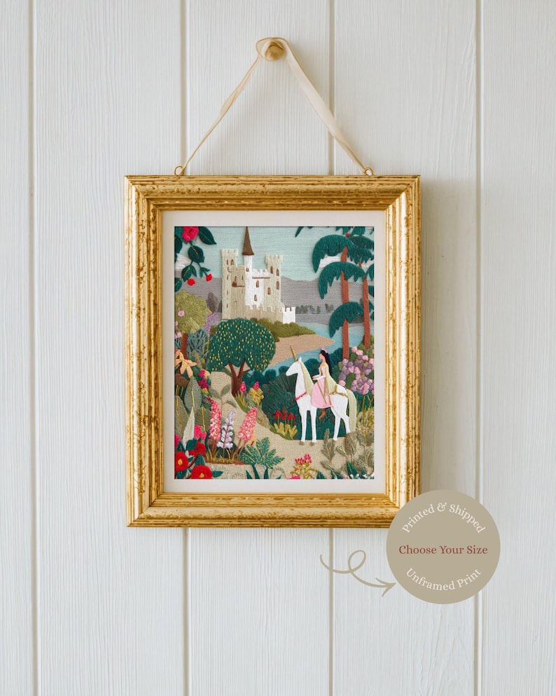 May include: A framed print with a gold-colored frame, depicting a fairytale scene. The artwork features a castle, a person riding a unicorn, and lush greenery. The print is hanging on a white wall with a beige ribbon.