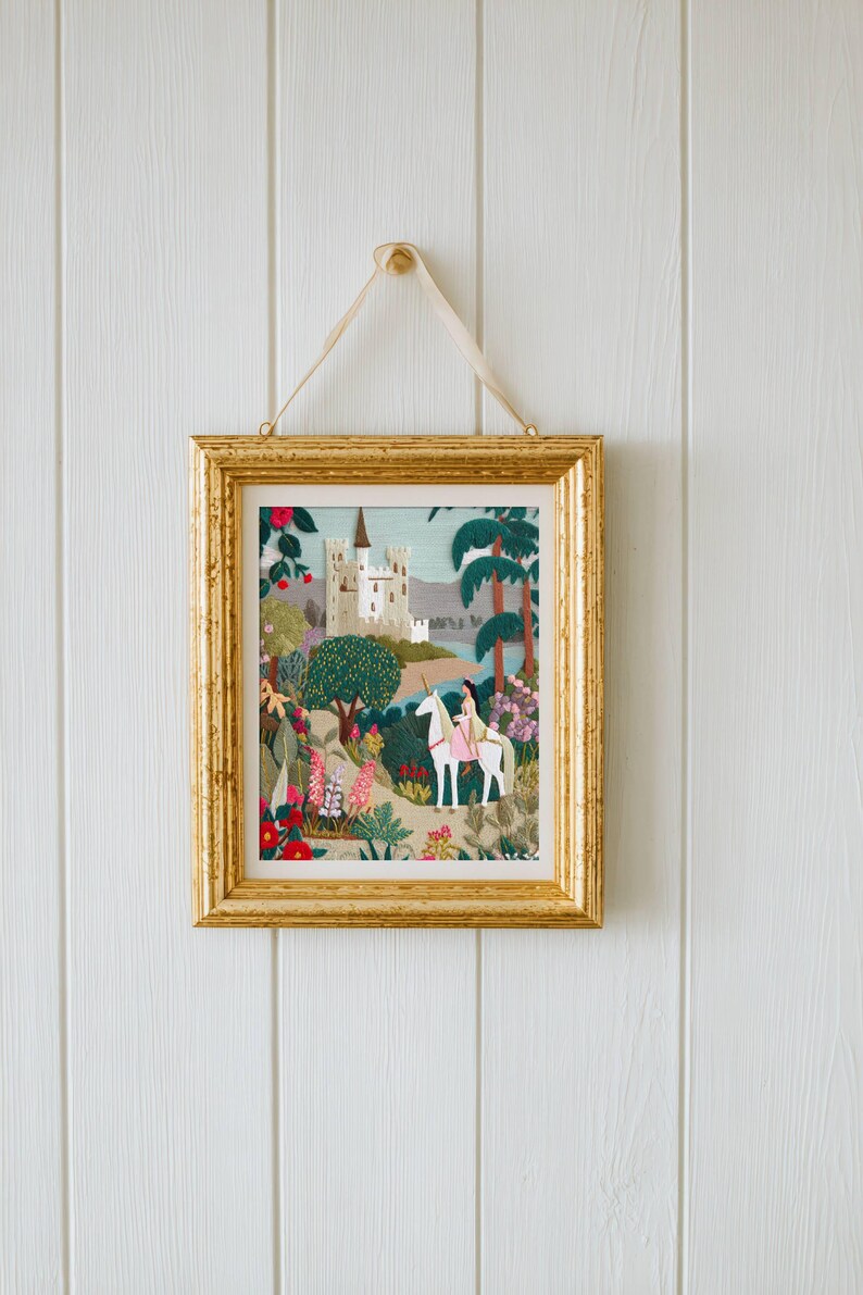 May include: A framed artwork with a gold-colored frame, hanging on a white wall. The artwork depicts a fairytale scene with a castle, trees, and a person riding a white horse. The artwork is colorful with pink, green, and blue hues.