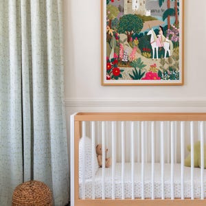 May include: A framed artwork depicts a fairytale scene with a castle, a person riding a unicorn, and lush gardens. The artwork hangs above a wooden crib with white slats and a patterned mattress. A woven basket and patterned curtains are also visible.