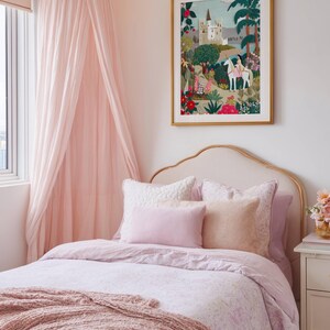 May include: A bedroom with a pink and gold theme. The bed has a light pink comforter, pillows, and a knitted throw. A framed picture with a castle scene hangs above the bed. A pink canopy and string lights add to the decor.