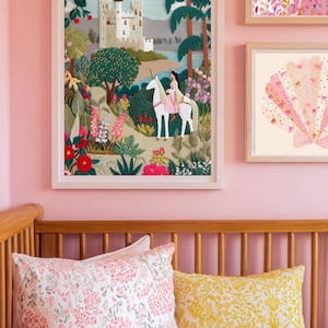 May include: Framed artwork featuring a fairytale scene with a castle, trees, and a person riding a unicorn. The artwork is in a white frame and hangs on a pink wall. Other framed art pieces are also visible. A wooden crib with floral pillows is in the foreground.