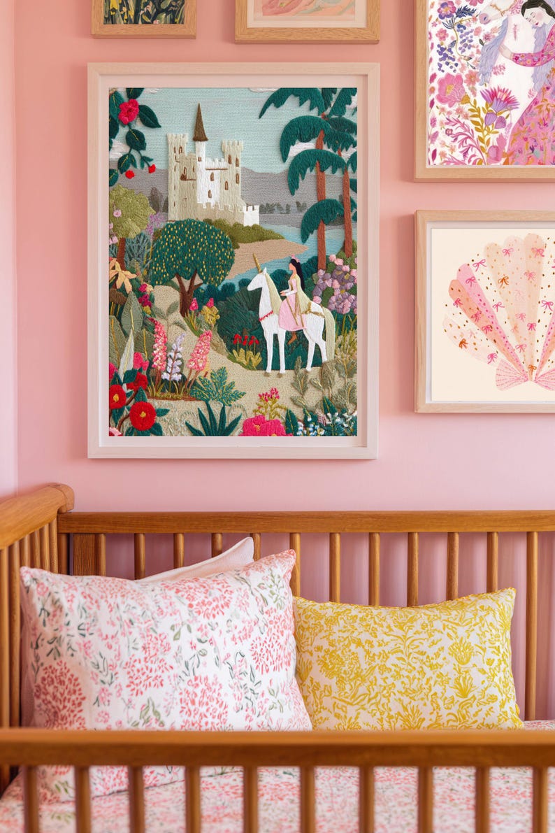 May include: Framed artwork featuring a fairytale scene with a castle, trees, and a person riding a unicorn. The artwork is in a white frame and hangs on a pink wall. Other framed art pieces are also visible. A wooden crib with floral pillows is in the foreground.