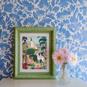 May include: A framed artwork with a green border depicts a fairytale scene with a castle, a person riding a white unicorn, and colorful flowers. The artwork is set against a blue and white floral wallpaper, with a vase of pink flowers in the foreground.