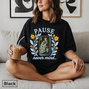 May include: A black t-shirt featuring a graphic print with the text PAUSE never mind. The design includes a central rectangular tile with the number 1 and a peacock illustration, surrounded by light blue leafy branches and orange flowers.