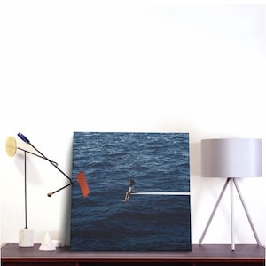 May include: A canvas print depicting a person sitting on a white diving board extending over a vast, dark blue ocean. The artwork is displayed on a dark wooden surface, alongside a modern lamp and abstract sculpture.