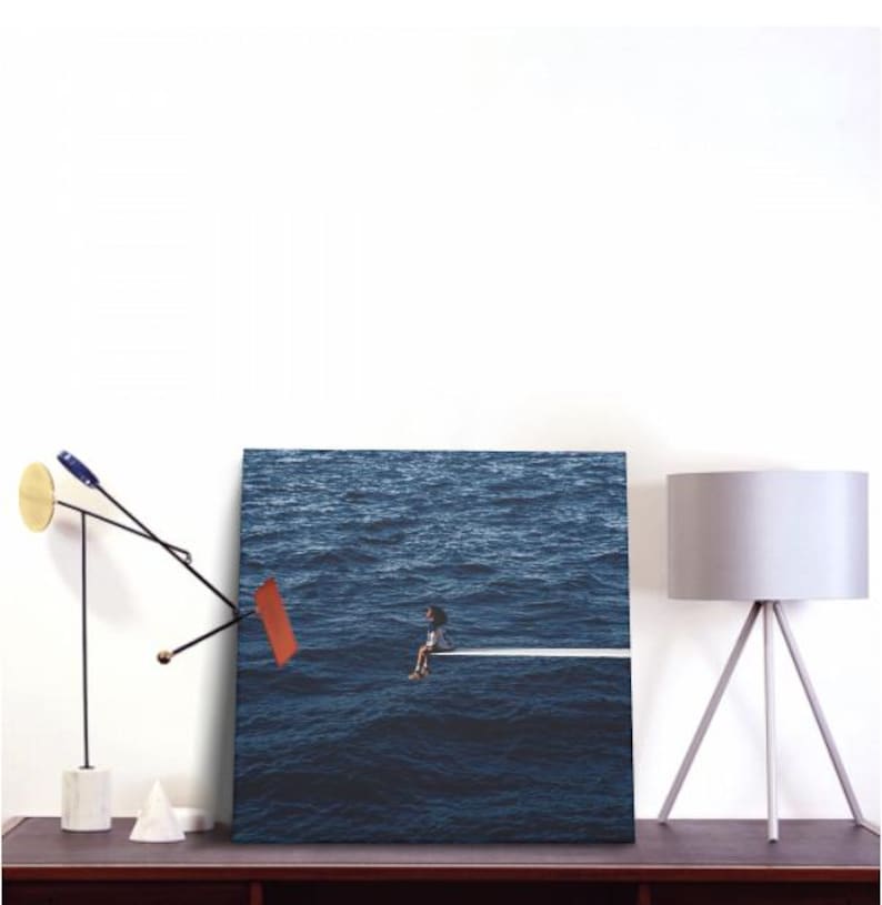 May include: A canvas print depicting a person sitting on a white diving board extending over a vast, dark blue ocean. The artwork is displayed on a dark wooden surface, alongside a modern lamp and abstract sculpture.