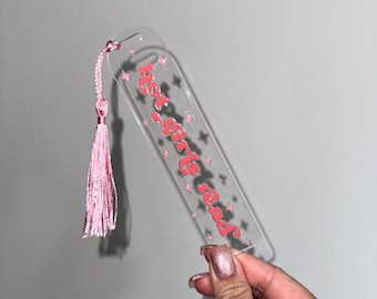 Peut inclure: Un marque-page en acrylique transparent avec un pompon rose. Le marque-page porte le texte "Hot girls read" en lettres roses avec des étoiles roses éparpillées autour du texte.