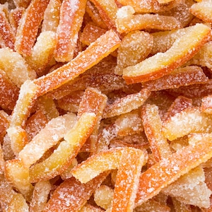 May include: Close-up of candied orange peel pieces. The peel is cut into strips and coated in granulated sugar. The colors range from light yellow to deep orange, with a dusting of white sugar crystals.