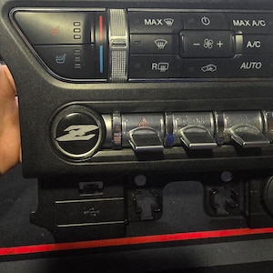 Puede incluir: Un salpicadero de coche negro con un panel de control plateado que incluye botones para el control del clima, el audio y otras funciones. El panel incluye etiquetas como "MAX", "A/C", "AUTO" y "R".