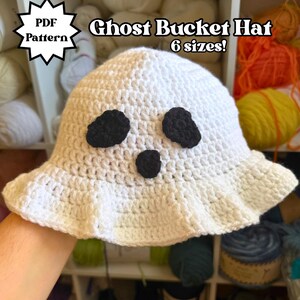 May include: A white crocheted bucket hat featuring two black oval eyes and a small black circular mouth. The text on the image reads PDF Pattern Ghost Bucket Hat 6 sizes!