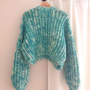 May include: A teal and white crocheted sweater with a cropped fit and long sleeves. The sweater has a loose, relaxed fit and a slightly oversized look.