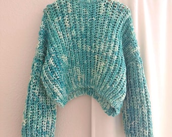 May include: A cropped, long-sleeved sweater featuring a chunky, ribbed knit texture. The garment is crafted from variegated yarn in shades of turquoise, teal, and white, creating a marbled appearance throughout the fabric with a rounded neckline.