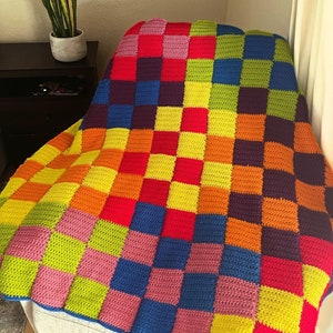 May include: A handmade crochet blanket featuring a vibrant checkerboard pattern. The textile is composed of numerous square blocks in bright shades of red, orange, yellow, lime green, blue, purple, and pink, arranged in a repeating grid layout.
