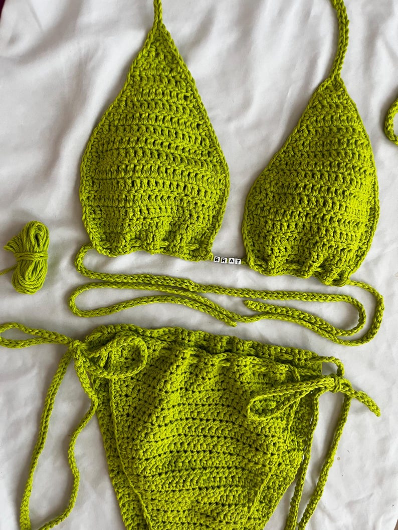 May include: A lime green crochet bikini top and bottom with matching ties. The top has a triangle shape and the bottom is a high-waisted style. The word "BRAT" is spelled out in black beads on the top.