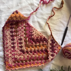 Puede incluir: Top de ganchillo hecho a mano en tonos rosa, naranja y marrón. El top presenta un patrón de granny square y lazos largos trenzados. También se ven un ganchillo y un ovillo de lana.